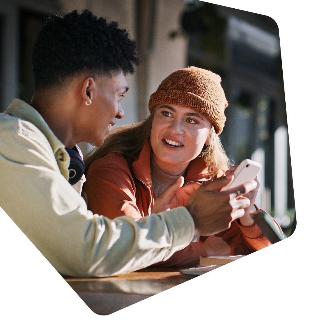 Zwei junge Menschen sitzen in einem Café und reden miteinander. Einer der beiden hält sein Handy in der Hand und zeigt etwas darin.
